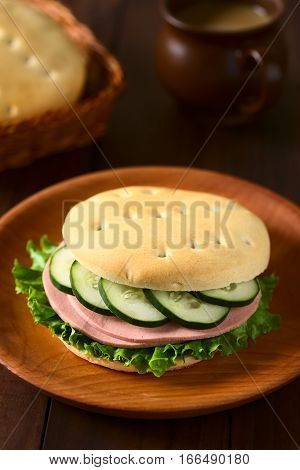 Traditional Chilean Hallulla bread roll prepared as a sandwich with lettuce cold cut and cucumber served on wooden plate photographed with natural light (Selective Focus Focus on the front of the sandwich)