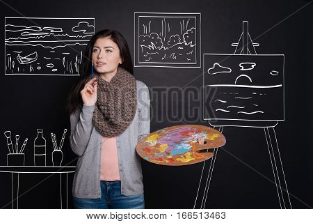 Work with nspiration. Pleasant thoughtful woman holding brush and thinking while enjoying painting