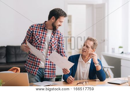 Discussion of the project. Nice diligent bearded man showing two sheets of paper to his colleague and discussing his ideas with him while working on the project together