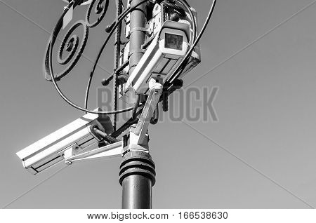 Security camera surveillance on the urban street lamppost on a background of the sky. Black and white image. Big Brother is watching through camcorder lens.