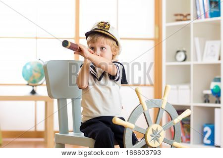 Kid dressed as a captain or sailor plays on chair as ship in his room. Child looks through telescope.