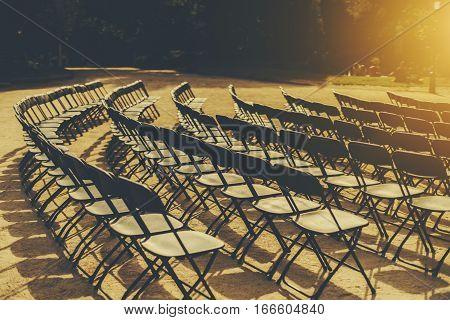 Empty seat rows of folding chairs on ground before a event parallel and rounded arranged multiple black chairs on street on sunny day in park with people on grass meadow in distance