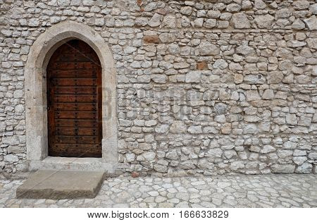 Wooden backdoor with pointed arch (gothic). The door is located in a wall with white irregularly shaped stone blocks which belongs to an ancient fortress or old castle. It is made of riveted wood planks and is locked and closed.
