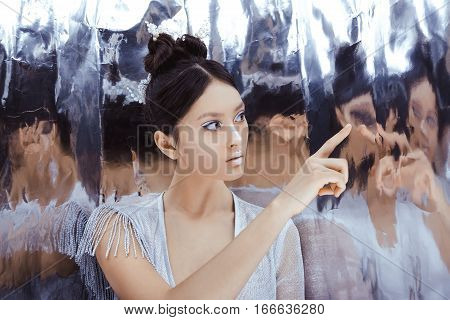 Portrait of futuristic young woman touching reflection in mirror. Reflection of our mind and soul concept. Beautiful young multi-racial asian caucasian model cyber girl in silver urban clothes with conceptual hairstyle and make-up in metallik silver capsu