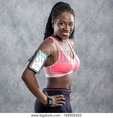 Close up portrait of attractive afro american female teen athlete in sportswear. Young woman wearing armband with smart phone and smart watch for activity tracking. Isolated against grey backdrop.