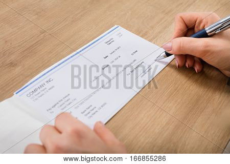 Close-up Of A Person Hand Signing Cheque At Wooden Desk