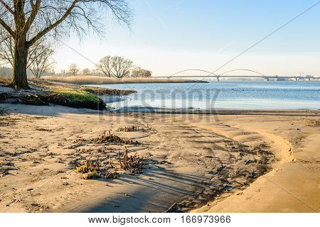 Small sandy beach along a wide Dutch river. It is in the late afternoon of a sunny day in the winter season. In the background a large arch bridge is visible.