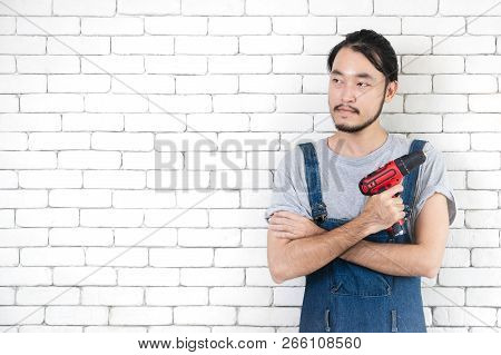 Young Asian Man Holding Power Drill Standing In Front Of White Brick Wall, Smiling And Looking At Ca