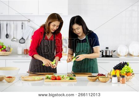 Beautiful Asian Woman In Red Shirt And Black  Apron Teaching Her Daughter How To Prepare Vegetables 