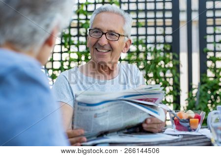 Senior couple reading newspaper during breakfast. Portrait of happy old man with grey hair reading the news with wife in the morning. Elderly couple discussing while reading newspaper outdoor.