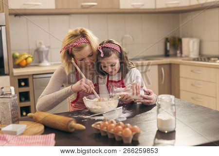 Beautiful Mother And Daughter Having Fun In The Kitchen; Daughter Pouring Milk While Mother Is Knead