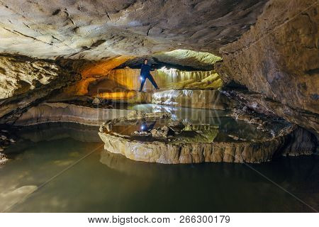 Man Tourist  Speleologist In Wetsuit With Flashlight In Beautiful Natural Cave With Cascades Of Unde