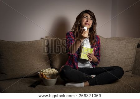 Beautiful Young Woman Sitting In The Dark On A Living Room Couch, Watching A Soap Opera And Crying