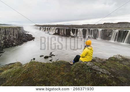 Woman In Yellow Raincoat Sitting Standing On The Cliff Edge Next To Selfoss Waterfall In Vatnayokull