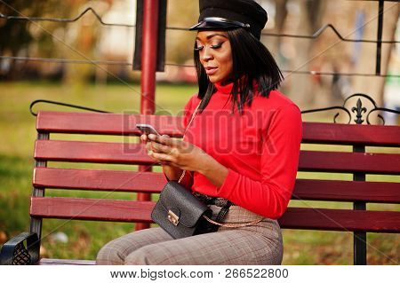 African American Fashion Girl In Newsboy Cap, With Handbag Posed At Street, Sitting On Bench With Mo