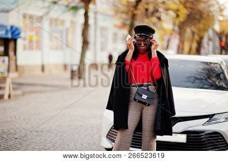 African American Fashion Girl In Coat, Newsboy Cap And Sunglasses Posed At Street Against White Busi