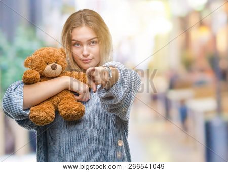 Young caucasian woman holding cute teddy bear over isolated background pointing with finger to the camera and to you, hand sign, positive and confident gesture from the front