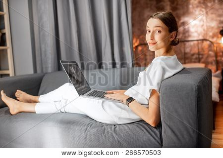 Young Woman In White Clothes Sitting On The Couch With Laptop At The Living Room Of The Modern Loft 