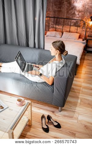 Young Woman In White Clothes Sitting On The Couch With Laptop At The Living Room Of The Modern Loft 
