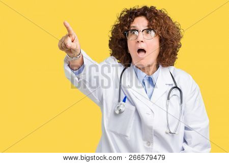 Middle ager senior doctor woman over isolated background Pointing with finger surprised ahead, open mouth amazed expression, something in front