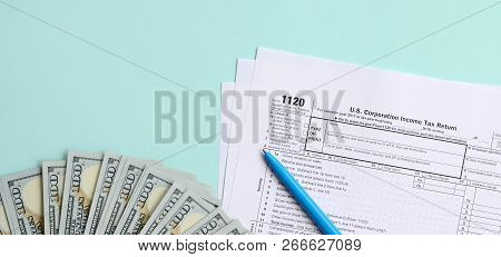 1120 Tax Form Lies Near Hundred Dollar Bills And Blue Pen On A Light Blue Background. Us Corporation