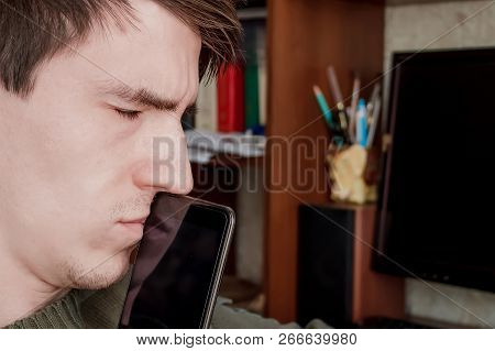 Thoughtful Guy With A Smartphone At His Mouth, With His Eyes Closed, Sits At The Desk