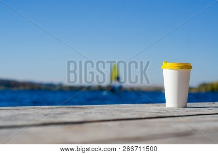 White Paper Cup With A Yellow Plastic Lid Stands On The Boards On The Shore Of The Lake