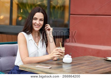Cheerful European Woman Enjoys Leisure Time, Drinks Milk Shake At Outdoor Cafeteria, Spends Weekened