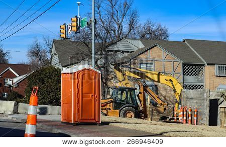 Portable Bathroom At Construction Site At Intersectio Of Urban Roads With Backhoe In Background And 
