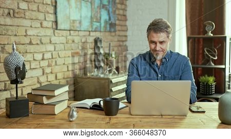 Older man tele working with laptop computer at home office in living room, thinking, looking busy.