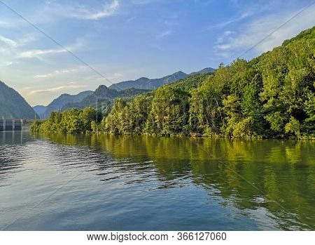 River Flowing Peacefuly With Green Trees On The Banks And Mountain Landscape In The Distant Backgrou
