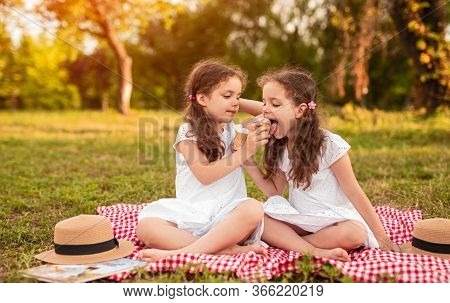 Little Girl Sitting On Checkered Blanket And Sharing Tasty Ice Cream With Twin Sister During Picnic 