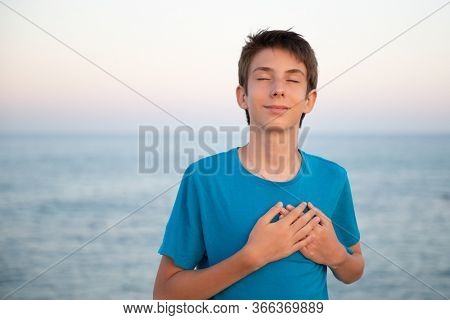 Handsome young happy boy with eyes closed at beach holds hands on chest, sunrise or sunset. Beautiful calm teen boy feels grateful, prays, thanks God, hopes, wishes dream come true, at sea coast. 