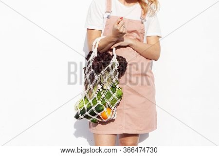 Young Woman In Light Summer Clothes Is Holding Mesh Shopping Bag With Vegetables, Greens Without Pla