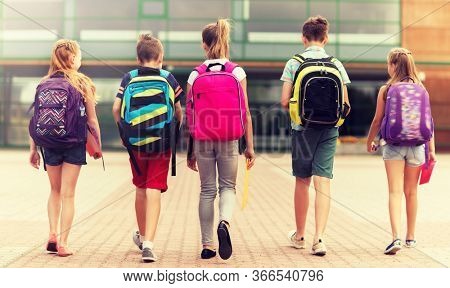 primary education, friendship, childhood and people concept - group of happy elementary school students with backpacks walking outdoors from back