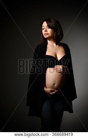 Brunette Pregnant Woman In Black Shawl, Pants And Bra, Bare Belly. She Looking Aside, Posing Sideway