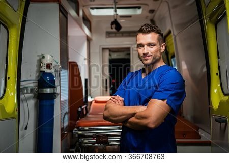 Handsome Paramedic In A Medical Uniform Looking At The Camera And Smiling, Ambulance Car In The Back