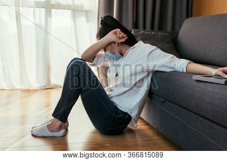 Grieving Young Woman In Medical Mask Obscuring Face With Hand While Sitting On Floor And Touching Ph