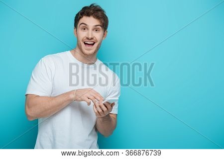Excited Guy Looking At Camera While Chatting On Smartphone On Blue Background