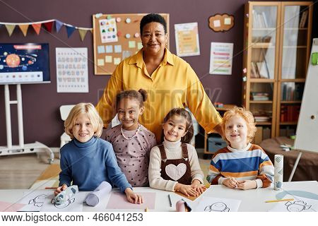 Happy Mature Teacher And Group Of Intercultural Little Learners Standing By Desk And Looking At Came
