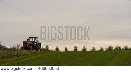 An Old Tractor Stands In A Field On Top Of A Hill. A Field With Growing Winter Grain. A Rather Dilap