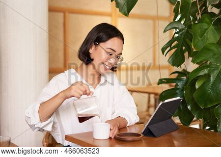 Freelance And Remote Workers. Smiling Young Woman Pouring Coffee In A Cup, Sitting In Cafe And Looki