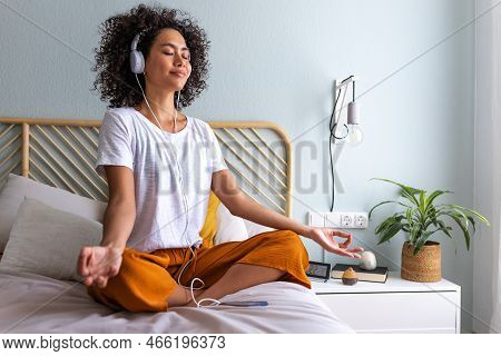 Young Woman Meditating At Home Sitting On Bed Following Online Meditation, Listening With Headphones