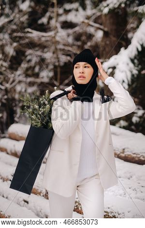 A Girl In A White Suit And Balaclava With A Package Of Christmas Trees In The Winter Forest On New Y