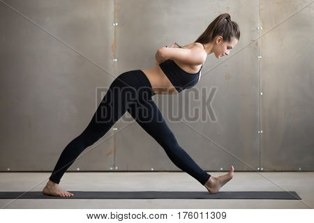 Young attractive woman practicing yoga, standing in One Sided Fold exercise, Ardha Parsvottanasana, Half Pyramid pose, working out, wearing black sportswear, cool urban style, full length, grey studio