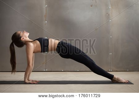 Young attractive smiling woman practicing yoga, standing in Upward Plank exercise, Purvottanasana pose, working out, wearing black sportswear, cool urban style, full length, grey studio background