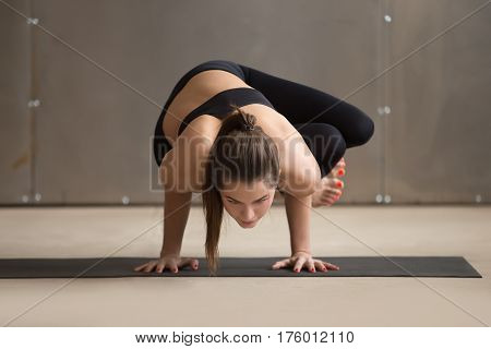 Young attractive yogi woman practicing yoga, standing in Parsva Bakasana exercise, Side Crane pose, working out, wearing black sportswear, cool urban style, full length, grey studio background