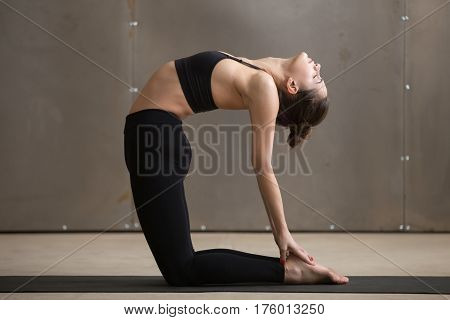 Young attractive yogi woman practicing yoga, doing Ustrasana exercise, Camel pose, working out, wearing black sportswear, cool urban style, full length, grey studio background, side view