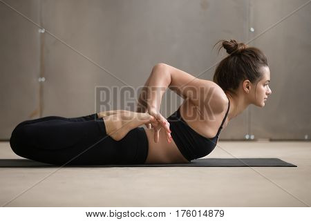 Young attractive yogi woman practicing yoga, stretching in Bhekasana exercise, Frog pose, working out, wearing black sportswear, cool urban style, full length, grey studio background, side view