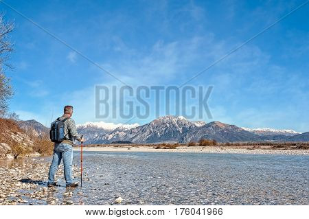 Mature Hiker On The Bank Of A River. Walking Toward Mountain.